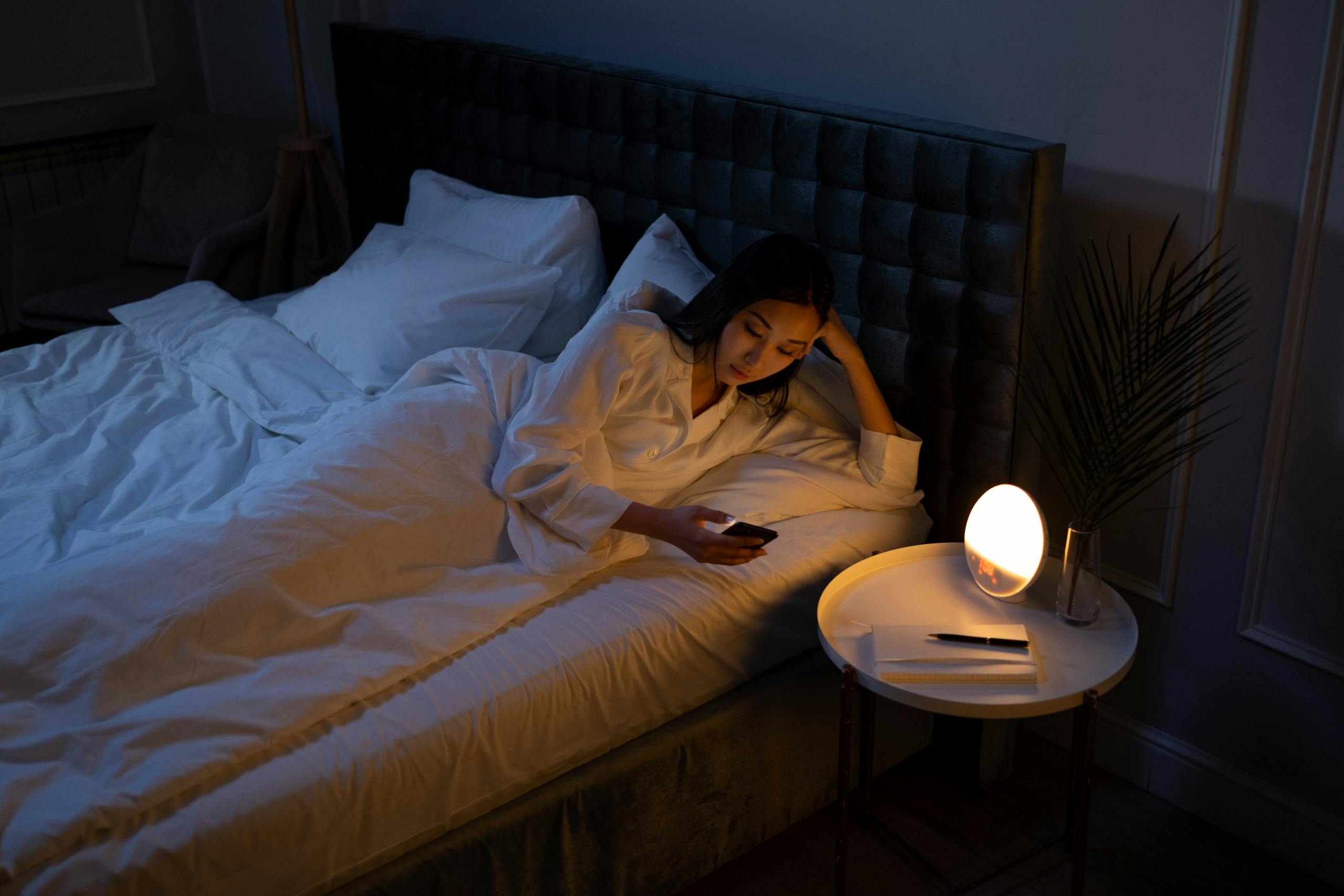 Finding Balance: How Acupuncture and Chinese Herbal Medicine Can Help with Anxiety and Depression 10 An Asian woman on a bed using her phone in a dim bedroom, reflecting on insomnia or night routines.